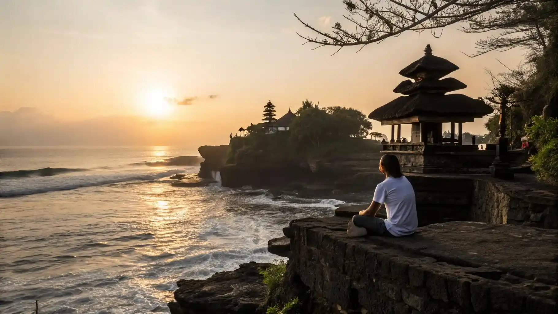 A visitor sitting above the sea temple of Tanah Lot as the sun sets